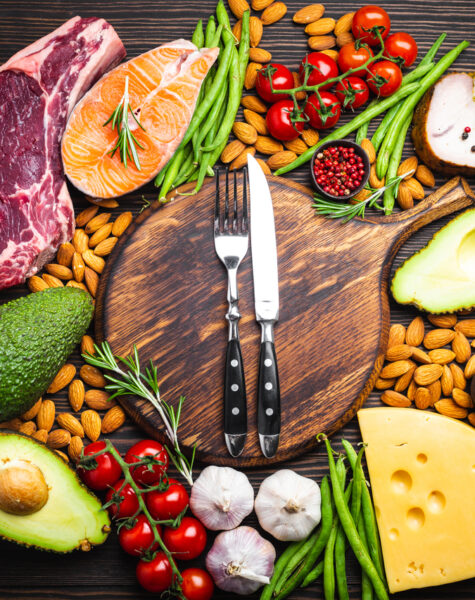 Knife and fork over wooden cutting board and ketogenic low carbs ingredients for healthy eating concept and weight loss, top view. Keto foods: meat, fish, avocado, cheese, vegetables, nuts
